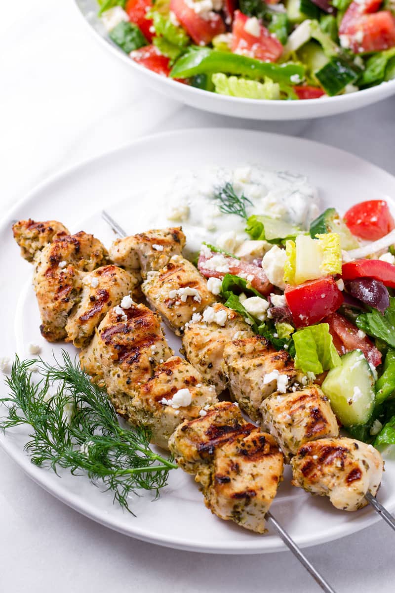 Two Greek style chicken kebabs on a plate with side of Greek salad and tzatziki sauce