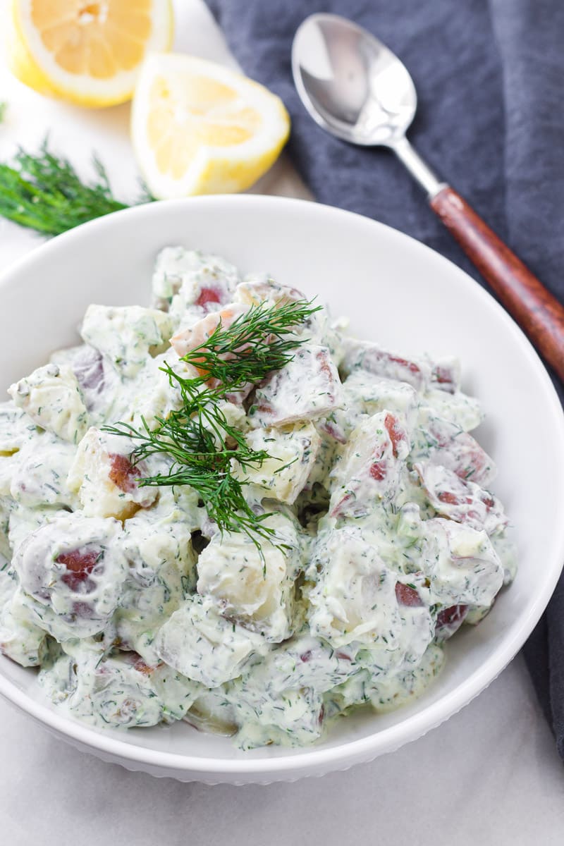 White bowl with creamy potato dill salad with a blue napkin and a wooden spoon in the back