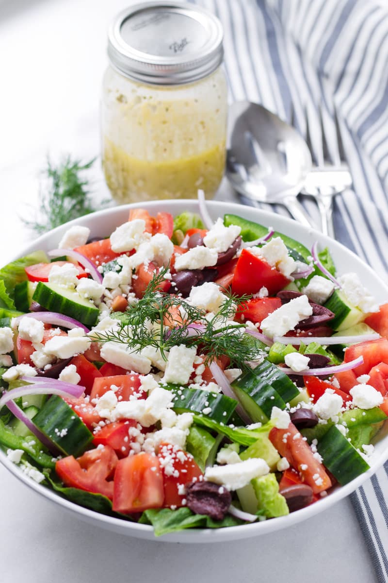 Greek salad on a white bowl and a mason jar halfway filled with dressing