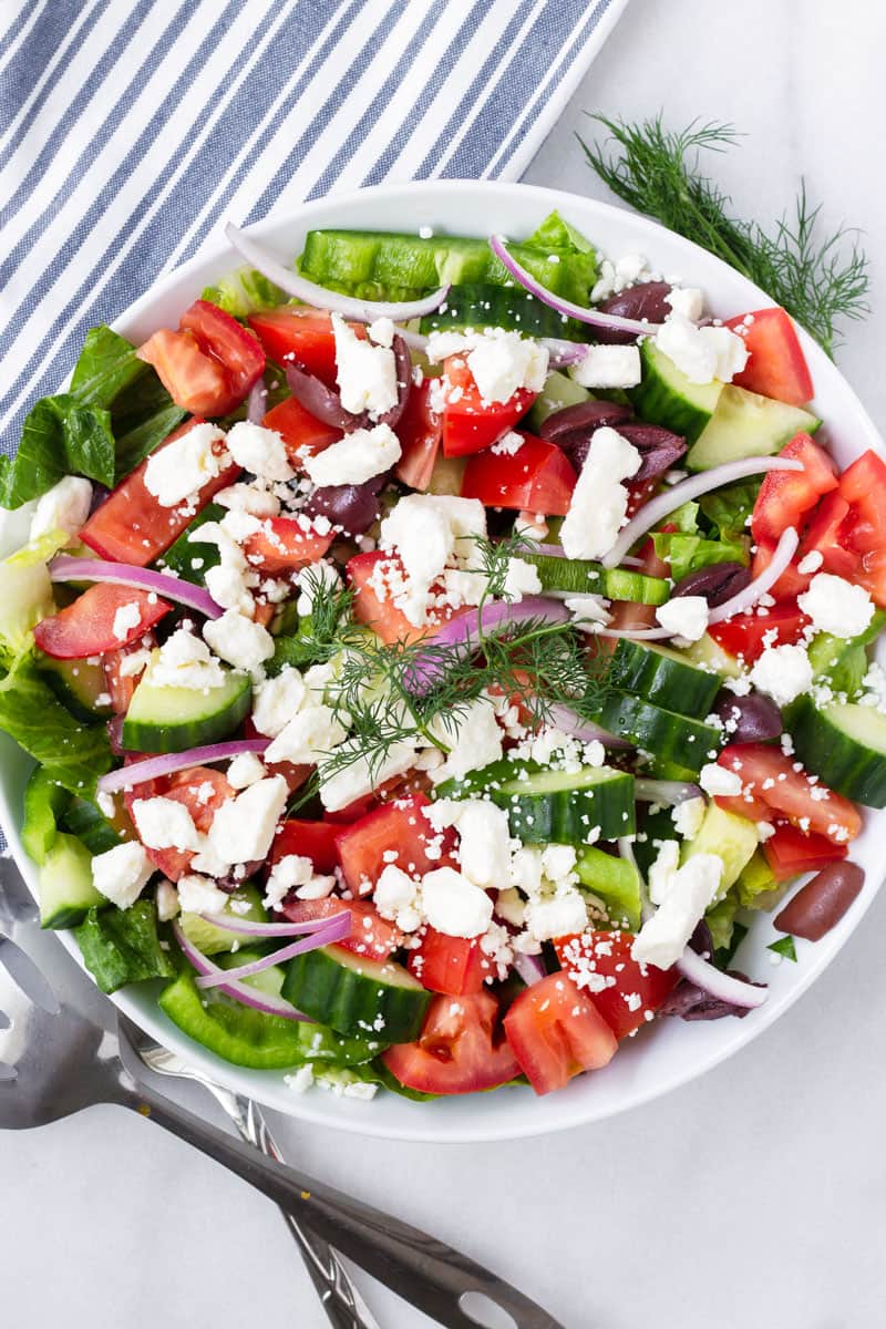 bowl with Greek salad on a striped white and blue napkin and serving spoons