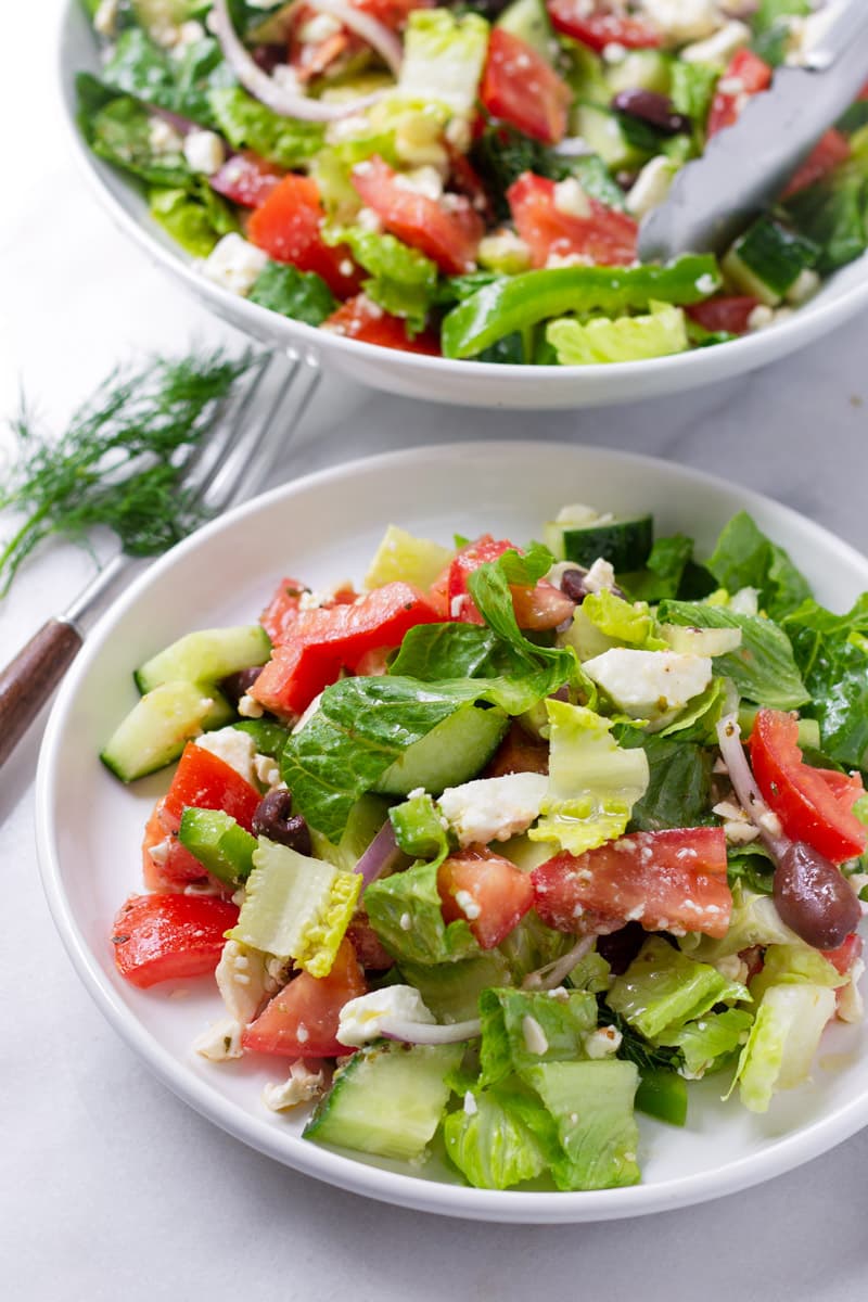 A small white plate with Greek Salad and a bowl of Greek salad in the background