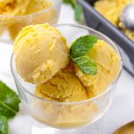 A glass bowl with three scoops of mango ice cream and mint garnish