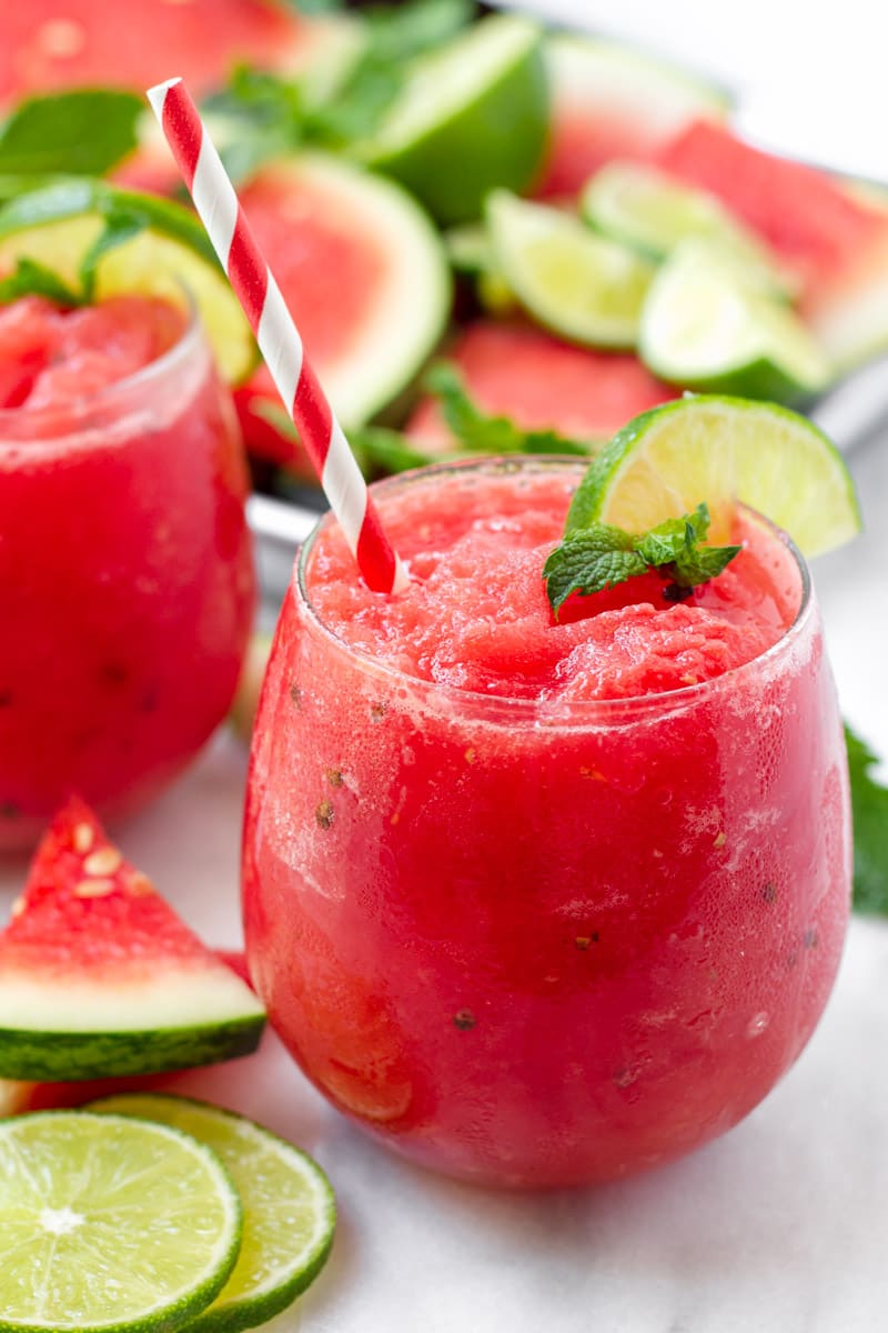 Red watermelon slush in a glass with red and white paper straw, plus lime and mint garnish