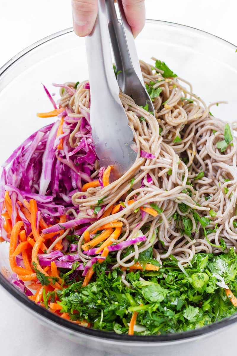 Bowl with noodle salad ingredients being tossed with tongs