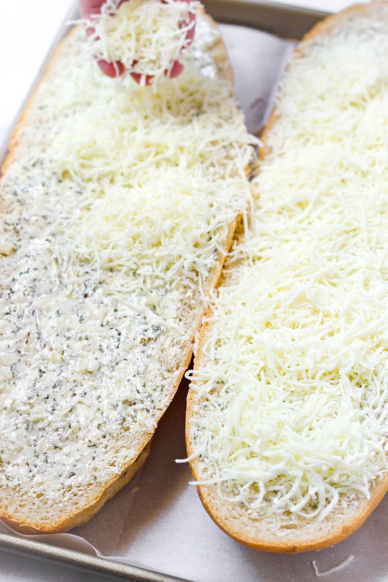 A loaf of bread split in half, spread with compound butter and topped with shredded cheese