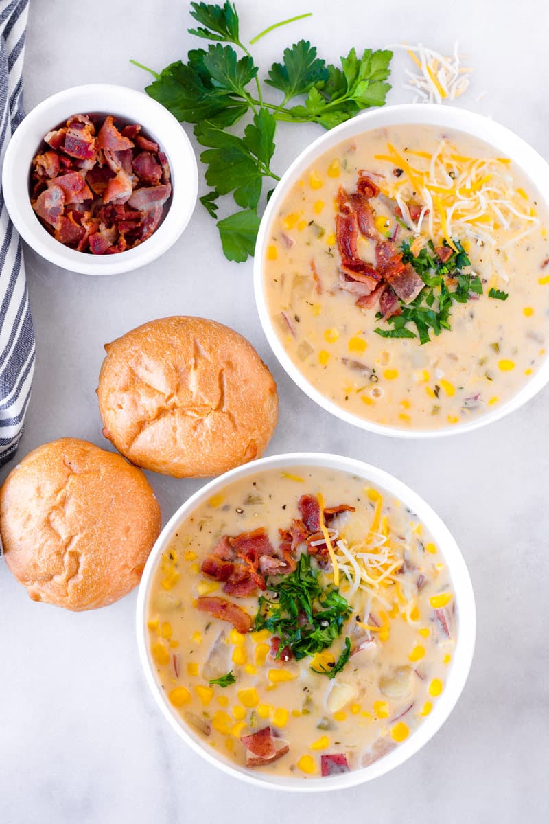 Top view of two bowls of potato corn chowder, two bread rolls, and bacon bits