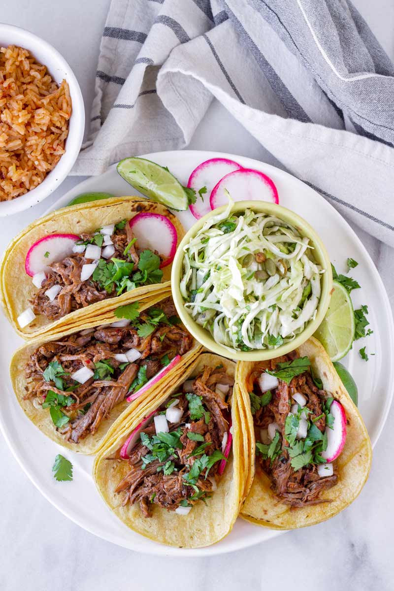 top view of four barbacoa tacos with radish and cilantro garnish, and served with rice and slaw