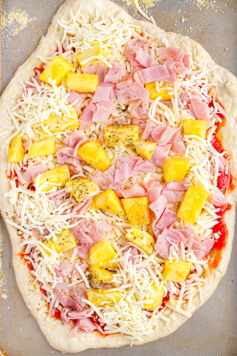 unbaked cheese pizza with toppings like pineapple, ham and oregano