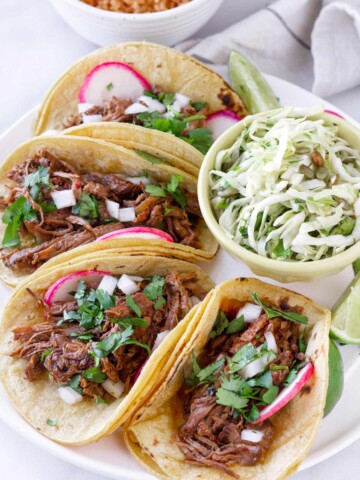 four beef barbacoa tacos arranged on a plate with a side of slaw
