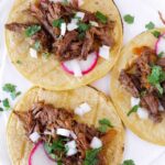top view of three corn tortillas with barbacoa beef, radishes, cilantro and onions