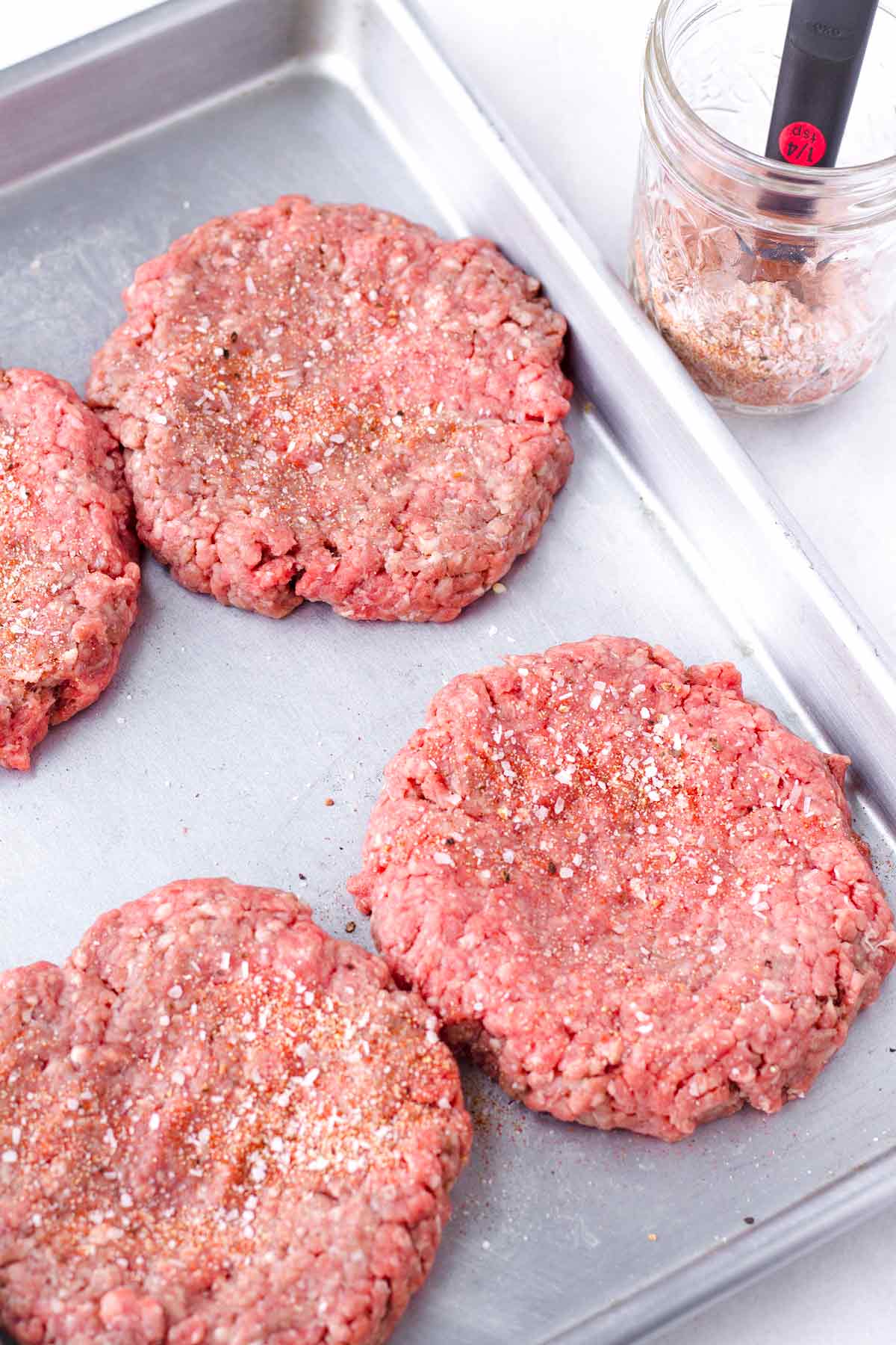 four seasoned meat patties on a sheet pan, next to a mason jar with seasoning