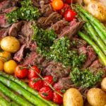 wooden platter with sliced skirt steak, chimichurri sauce, and grilled vegetables