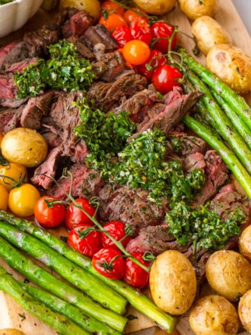 wooden platter with sliced skirt steak, chimichurri sauce, and grilled vegetables