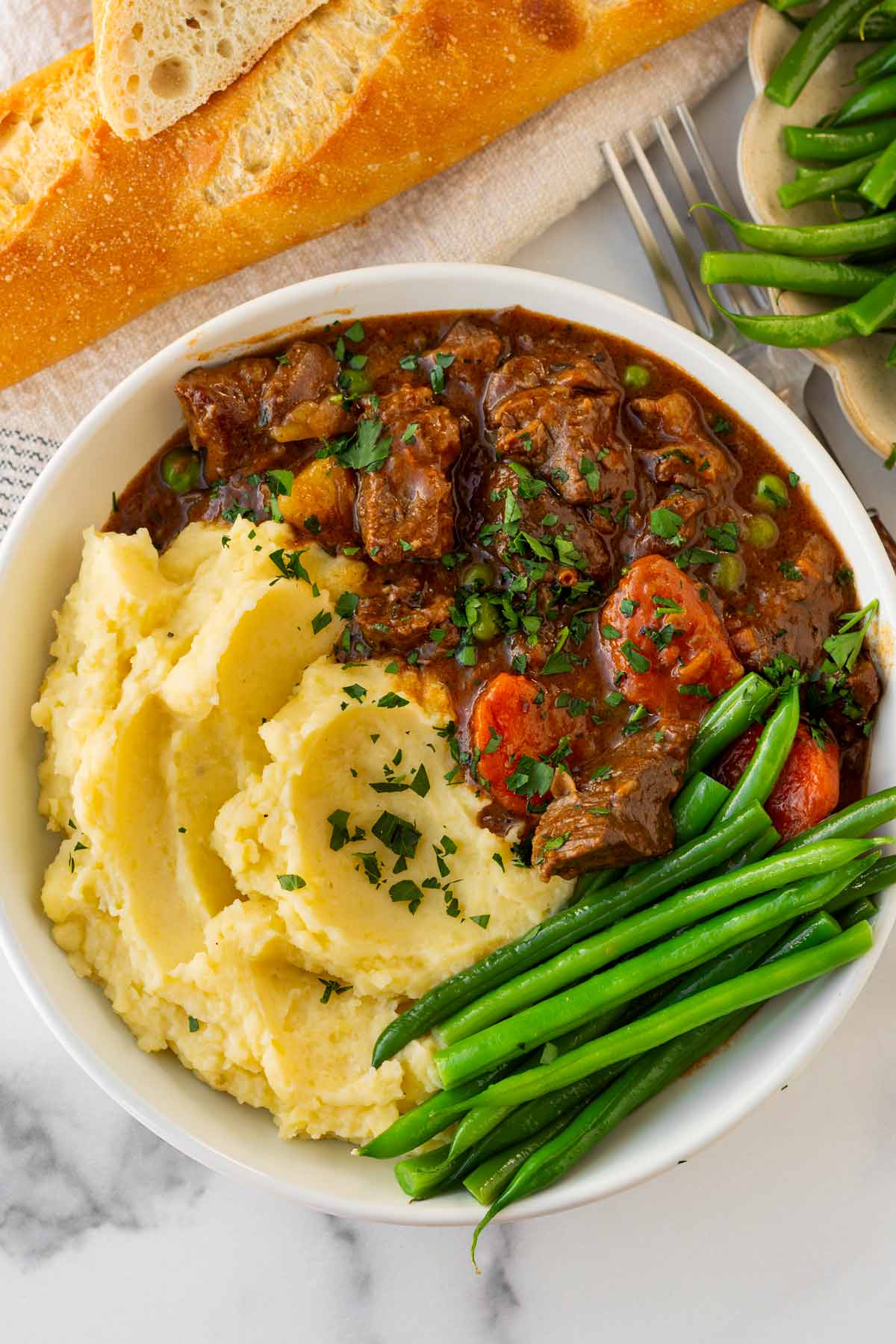 bowl of beef stew cooked in the oven served over mashed potatoes and a side of green beans