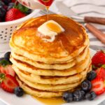 drizzling maple syrup over a stack of six buttermilk pancakes